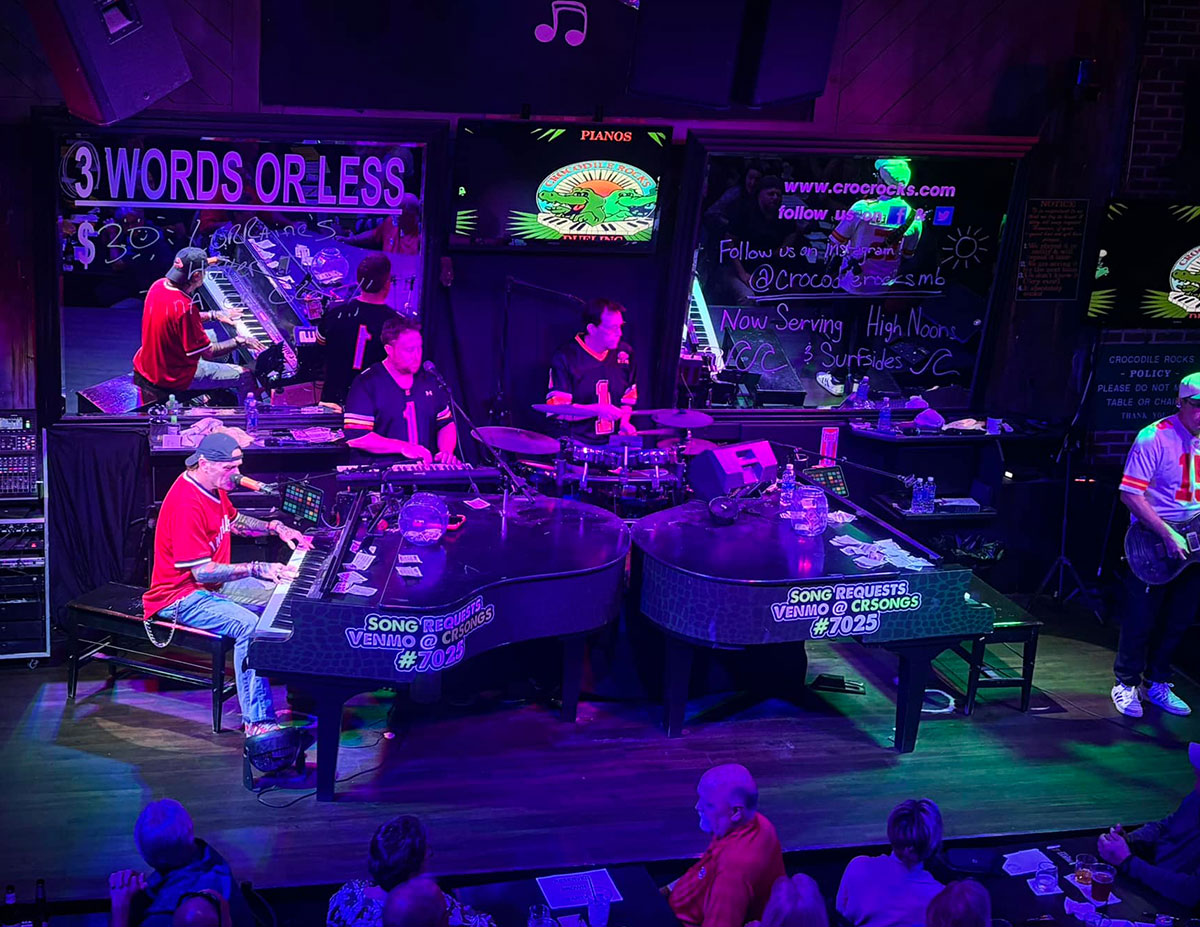 Two grand pianos on a stage with musicians performing for a crowd
